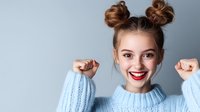 Young Caucasian woman with double bun hairstyle wearing light blue sweater expressing joy and excitement with raised fists against gray background.