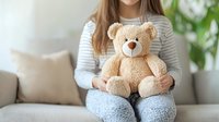 Young woman in casual wear holding classic beige teddy bear while sitting on gray couch at home, selective focus on plush toy, cozy atmosphere with green plants.