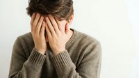 Young Caucasian woman covering face with hands showing stress, depression or headache in brown knit sweater against neutral background for mental health concepts.