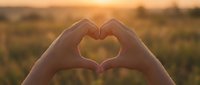 Hands forming a heart shape against a warm golden sunset over a meadow, symbolizing love, hope, and connection in nature.