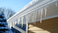 Crystalline icicles hanging from white roof gutter against blue winter sky, with snow-covered trees in soft focus background creating seasonal atmosphere.