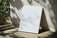 Blank white canvas mockup standing on wooden table with natural shadow overlay and plant decoration. Artistic setup for branding presentation.