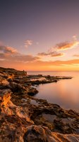 Dramatic coastal landscape at golden hour with rocky shoreline, calm ocean waters, and vibrant sunset sky featuring orange and purple hues. Perfect for travel and nature projects.