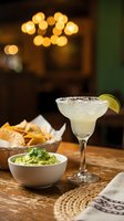 Margarita cocktail with salt rim and lime garnish served with tortilla chips and guacamole dip on wooden table in warm bar setting.