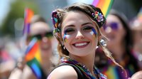 Young Caucasian woman with rainbow face paint and glitter makeup smiling at pride parade celebration, wearing colorful festival attire and flower hair accessories.