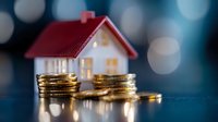Small red-roofed house model with stacked gold coins on blue background with bokeh lights, representing real estate investment, property finance and mortgage concepts.