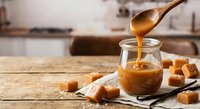 Golden caramel sauce dripping from wooden spoon into glass jar on kitchen counter with caramel candies scattered around for dessert preparation.