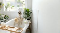 Skeleton relaxing in bathtub with plants and candles in bright bathroom interior for Halloween decoration or medical education concept.