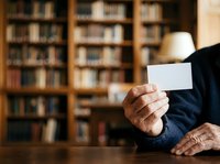 Mature man with wrinkled hands holding blank white card in library setting with wooden bookshelves background. Suitable for business, education, and professional concepts.