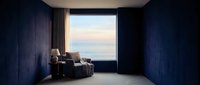 Luxury modern bedroom with dark blue walls, contemporary furniture, and expansive ocean view through floor-to-ceiling window at sunset. Interior design.