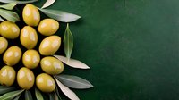 Fresh green olives arranged with olive leaves on dark green textured background, corner composition with copy space for food photography and menu design.