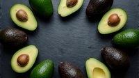 Fresh ripe avocados arranged in circle on dark slate background. Healthy organic fruit halves with pits showing creamy green flesh for cooking recipes.
