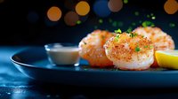 Pan seared sea scallops garnished with microgreens and lemon on dark plate with creamy sauce, dramatic lighting creates elegant restaurant atmosphere.
