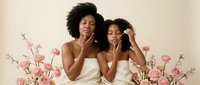Two women with dark curly hair and medium skin tone pose together surrounded by pink flowers against a neutral background. Both wear white sleeveless tops and display natural, joyful expressions. Suitable for beauty, wellness, and lifestyle marketing.