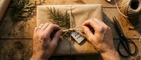Hands wrapping kraft paper gift box with twine and dried greenery, overhead view of eco-friendly present preparation for holidays or special occasions.