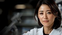 Young Asian female medical professional in white lab coat smiling confidently in clinical setting with blurred background, representing healthcare and medical expertise.