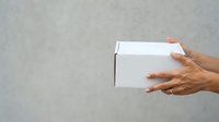Person with medium skin tone holding white cardboard box against neutral gray background, suitable for delivery, shipping, packaging, and e-commerce concepts.