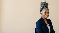 Mature African American woman with gray braided updo hairstyle wearing navy blazer and white shirt, smiling at camera against neutral beige background for corporate and professional use.