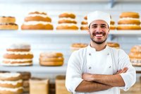 Hispanic male baker in white chef uniform smiling with crossed arms in front of freshly baked layered cakes displayed on bakery shelves, professional culinary setting.