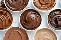Smooth swirls of melted chocolate in various shades from dark to milk and white displayed in white ceramic bowls, creating appetizing patterns and textures for dessert making.