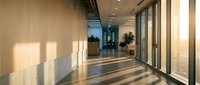 Modern office corridor with floor-to-ceiling windows and natural light creating geometric shadows on polished floors in contemporary corporate building.
