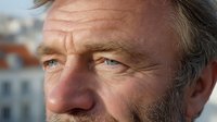 Close-up portrait of mature Caucasian man with blue eyes and stubble against blurred urban background, conveying wisdom and life experience.