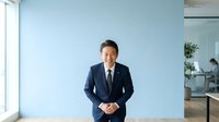 Asian male professional in dark business suit and tie standing in modern office with light blue wall, hands clasped, confident posture for corporate, leadership, and business portraits.