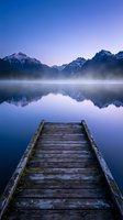 Wooden dock extending into misty mountain lake at dawn with snow-capped peaks reflected in calm water creating serene landscape for meditation and relaxation.
