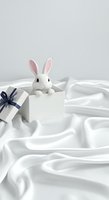 White rabbit toy sitting in gift box with blue ribbon on white silk fabric background, suitable for Easter, holiday greeting cards, and celebration designs.