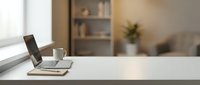 Clean white desk workspace with laptop computer and coffee cup in modern home office interior with blurred background shelving and natural window light.