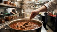 Chef stirring rich tomato meat stew in large pot in busy professional restaurant kitchen with copper cookware and steam rising.
