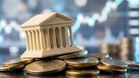 Miniature bank building with classical columns sits on pile of golden coins against blurred financial chart background representing banking and investment.