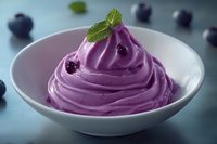 Creamy purple blueberry mousse dessert topped with fresh mint leaf and whole berries served in white ceramic bowl on blue background.