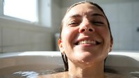 Young woman of mixed ethnicity with dark hair pulled back, smiling broadly while relaxing in a bathtub filled with water. Natural bathroom setting with white tiles and window light. Wellness and self-care concept.