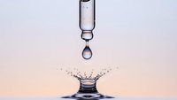 Water droplet falling from glass pipette creating splash crown on surface with soft gradient background for scientific research concepts.