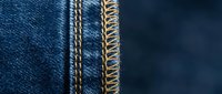 Close-up macro shot of blue denim fabric with golden yellow contrast stitching and frayed edges, showcasing textile craftsmanship and garment construction details.