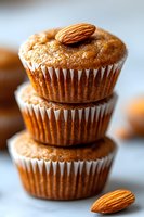 Stack of fresh almond muffins with whole nuts on top, showcasing golden-brown color and paper liners against light background, macro food photography.