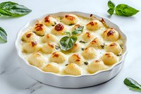 Homemade potato gratin dauphinois in white ceramic baking dish garnished with fresh basil leaves on marble surface, traditional French cuisine.