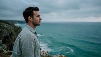 Young adult male with dark hair and light skin, wearing a gray linen shirt, gazing toward the ocean while standing on a rocky coastal cliff. Suitable for travel, lifestyle, and contemplation themes.