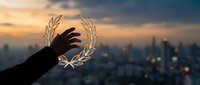 Silhouetted hand holding glowing laurel wreath against urban skyline at sunset, symbolizing achievement, success, and victory in business and personal goals.