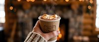 Hand holding a steaming hot cocoa cup with marshmallows by a cozy fireplace, warm winter holiday atmosphere, comfort drink concept.