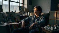 Young male artist sitting in messy creative studio workspace surrounded by art supplies, canvases and natural window light for artistic inspiration.