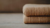 Neatly folded beige knitted sweaters showing detailed wool texture and stitching pattern on neutral background, soft focus creates cozy autumn mood.