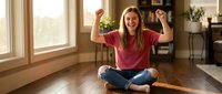 Young woman celebrating success sitting cross-legged on wooden floor at home, raising arms in victory gesture with joyful expression in bright living room.