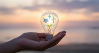 Hand holding glowing light bulb with renewable energy symbols solar panel and wind turbine inside at sunset representing sustainable innovation and green technology solutions.