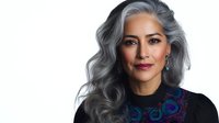 Confident mature Hispanic woman with silver gray hair wearing elegant black top with colorful embroidered neckline on white background isolated.