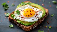 Sunny side up egg on multigrain toast with sliced avocado, red pepper flakes and fresh herbs. Healthy breakfast on dark background. Modern food photography.
