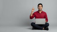Young man with dark hair and beard, South Asian ethnicity, wearing red shirt, sitting cross-legged with laptop, pointing upward with index finger, isolated on gray background for web and marketing use.
