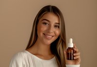 Young woman with long brown hair wearing white top holds brown glass dropper bottle against neutral beige background, displaying natural skincare product with genuine smile.