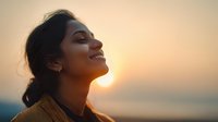Young South Asian woman smiling with eyes closed at sunset, golden light illuminating her profile against blurred ocean background, conveying peace and joy.